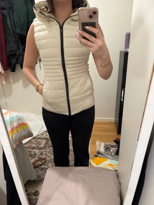 Beige dunväst från Zara med huva - Säljer en beige dunväst från Zara med huva och svarta detaljer. Västen har quiltad design, dragkedja framtill och ribbade partier på sidorna för extra komfort. Perfekt att slänga över en hoodie eller tröja för en chill look.