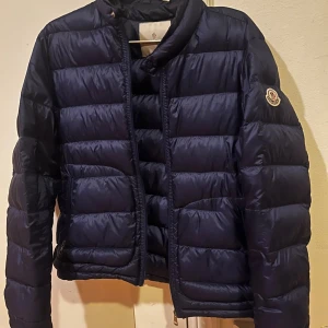 Mörkblå dunjacka från Moncler - Säljer en mörkblå dunjacka från Moncler med klassisk quiltad design och diskret logga på ärmen. Jackan har dragkedja framtill, ståkrage och två fickor med dragkedja. Perfekt för kyliga dagar och riktigt snygg till streetwear.