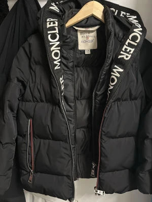 Svart Moncler pufferjacka barn - Säljer en svart pufferjacka från Moncler i barnstorlek 7-8 år. Jackan har huva med Moncler-logga, två fickor med dragkedja och coola detaljer i rött och vitt. Perfekt för kalla dagar och riktigt snygg streetstil. Namnet kan tvättas bort. 