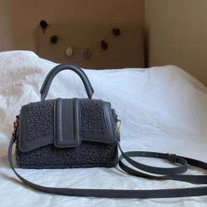 Mörkgrå axelväska i teddy-material - Säljer en mörkgrå axelväska från hm med handtag. Väskan är gjord i mjukt teddy-material med detaljer i syntetläder. Magnetlås framtill och stilren design som sticker ut. Bandet tillhör inte!!
