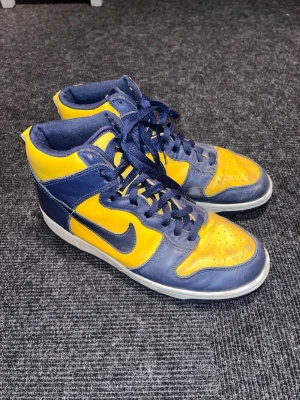 Nike Dunk Michigan  - Säljer ett par Nike Dunk High sneakers i blått och gult skinn med klassisk vit sula och blå snörning. Skorna har en hög siluett och tydlig Nike swoosh på sidan. Perfekta för dig som vill sticka ut med färg och retrostil.