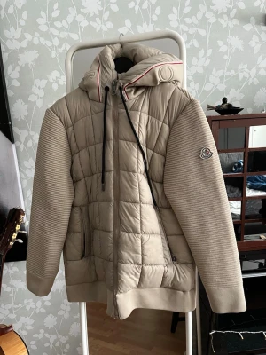 Beige pufferjacka från Moncler - Säljer en beige pufferjacka från Moncler med quiltad kropp och stickade ärmar. Jackan har huva med dragsko, diskret Moncler-logga på ärmen och dragkedja framtill. Snygg detalj med röd rand på huvan och ribbade muddar. Perfekt för kyliga dagar.
