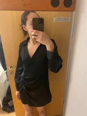 Svart omlottklänning från Zara - Säljer en svart kort omlottklänning från Zara med långa ärmar och krage. Klänningen har snygg drapering i sidan och en v-ringad urringning.  Snygg till middagar, fester och passar även en storlek S. Bra kvalité 