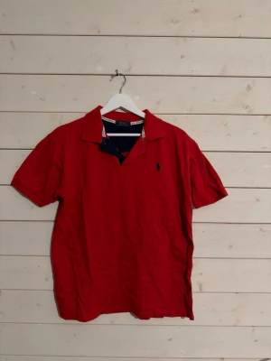 Röd pikétröja från Polo Ralph Lauren - Säljer en klassisk röd pikétröja från Polo Ralph Lauren med korta ärmar och krage. Tröjan har en liten broderad logga på bröstet och är tillverkad i mjuk bomull. Perfekt för en chill och stilren look.