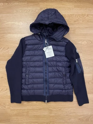 Mörkblå Moncler cardigan med huva - Säljer en mörkblå pufferjacka från Moncler med stickade ärmar och quiltad kropp. Jackan har huva, dragkedja framtill och en ficka med dragkedja på ärmen. Klassisk Moncler-logga på vänster ärm och coola detaljer som ribbade muddar. Det står att den är i Storlek 2xl men passar M/L