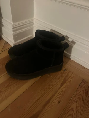 Svarta mockaboots med platt sula - Säljer ett par svarta uggs från din sko. Dom är använda en design och är i gott skick.