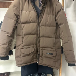 Brun dunjacka från Canada Goose s - Säljer en äkta  brun dunjacka från Canada Goose med huva och äkta päls detalj . Jackan har två stora fickor framtill med lock och dragkedja, samt ribbade muddar vid ärmsluten. Perfekt för kalla dagar och riktigt snygg till vinterlooken.