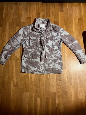 Camouflage jacka frön Samsøe Samsøe - Snygg vindjacka från Samsøe Samsøe i grått och beige camouflage-mönster. Jackan har flera stora fickor framtill, hög krage och dragkedja med knappslå. Tillverkad i slitstark bomull, perfekt för dig som gillar streetwear och utility-vibe.