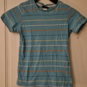 Blå randig t-shirt i bomull - Blå t-shirt med tunna horisontella ränder i vitt, orange, gult och mörkblått. Klassisk rund hals och korta ärmar. Tillverkad i mjuk bomull, perfekt för en avslappnad stil. En liten prick på framsidan (se bild) och en på baksidan (se bild) som inte går bort i tvätten. Dock inget man tänker på. Hund finns i hemmet. Kolla in mina andra annonser för samfrakt! 