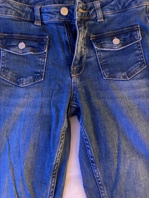 Blå jeans med ficklock och knappar - Säljer ett par klassiska blå jeans med framfickor som har lock och metallknappar. Jeansen har kontrastsömmar och är tillverkade i slitstarkt denimtyg. Passar dig som gillar en tidlös look med extra detaljer på fickorna. Sitter normal/medelhöjd i midjan. Storlek 164 men sitter fint på xs/s. Pris går att diskuteras💗