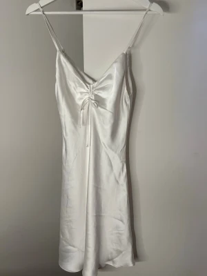 Vit kort satin klänning med snörning - Säljer en vit klänning i satin med smala axelband och  snörningsdetalj framtill. Klänningen är kort.