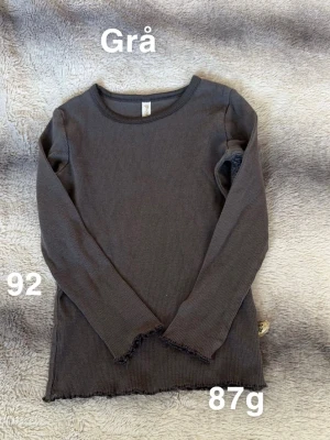 Grå ribbad långärmad topp - En enkel och stilren grå långärmad topp. Modellen har rund halsringning och vågiga kanter vid ärmslut och nederkant för en extra detalj. Perfekt att matcha med jeans eller kjol.🌸 Fint använt skick. Storlek 92.