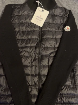 Svart quiltad jacka från Moncler - Säljer en svart quiltad jacka från Moncler med stickade ärmar och hög krage. Jackan har dragkedja framtill, två sidofickor och klassisk Moncler-logga på ärmen. Materialmix med blankt syntet på kroppen och stickad struktur på ärmarna ger en riktigt clean look.