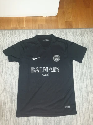 Svart PSG x Balmain Nike t-shirt S - Svart t-shirt från Nike med PSG och Balmain Paris-logga på bröstet. Tillverkad i Dri-FIT-material för extra komfort. Klassisk rund hals och kort ärm, perfekt för dig som gillar sportig och stilren look.