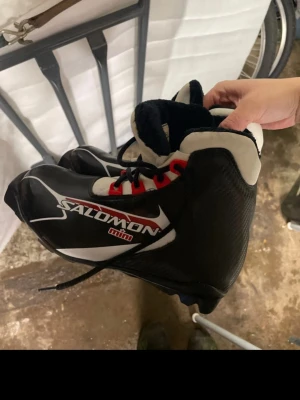 Salomon svarta skidpjäxor med röd snörning - Svarta skidpjäxor från Salomon med vita och röda detaljer samt röd snörning. Yttermaterialet är syntet och insidan är fodrad för extra värme. Sulan har SNS PROFIL-system för bra grepp och stabilitet. Perfekt för längdskidåkning under vintern.