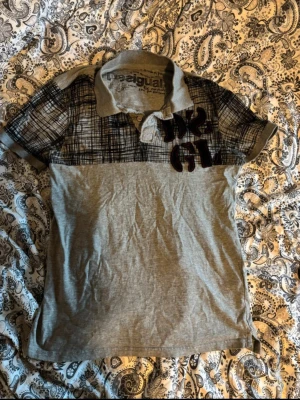 Grå mönstrad pikétröja Desigual - Säljer en unik pikétröja från Desigual i grått med svart rutigt mönster upptill och tryck på bröstet. Tröjan har klassisk krage, korta ärmar och knappar vid halsen. Tillverkad i mjuk bomull, perfekt för dig som gillar en lite annorlunda stil.