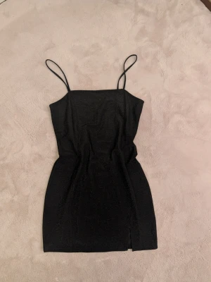 Svart glittrig kort klänning - Snygg svart klänning med glittrigt tyg och smala axelband. Modellen är kort och har en liten slits framtill. Perfekt för dig som gillar en stilren look med lite extra shimmer. Klänningen är ärmlös och har en figurnära passform.