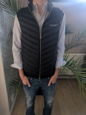 Svart dunväst från Armani Exchange - Snygg svart dunväst från Armani Exchange med diskret logga på bröstet. Västen har quiltad design, hög krage och dragkedja framtill. Insidan är ljusgrå och materialet är lätt och smidigt, perfekt att slänga över en skjorta eller hoodie. Sckicket är 10av10 envart använd fätal gånger