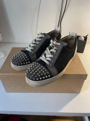 Christian Louboutin sneakers med nitar - Säljer ett par svarta och grå sneakers från Christian Louboutin med vita snören och silverfärgade nitar över tån. Dustbag, box och extra nitar ingår! Tveka inte att höra av dig vid frågor!