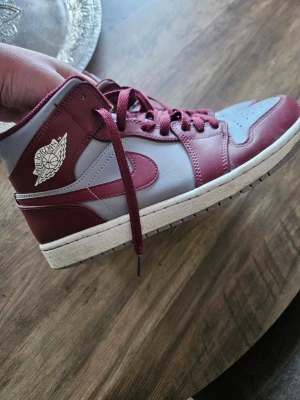 Nike Air Jordan 1 vinröd och grå - Säljer ett par Nike Air Jordan 1 i vinrött och grått skinn. Klassisk high-top modell med snörning, rund tå och vit sula. Swoosh-logga på sidan och Air Jordan-märket på ankeln. Perfekta för dig som vill sticka ut med stilrena sneakers.