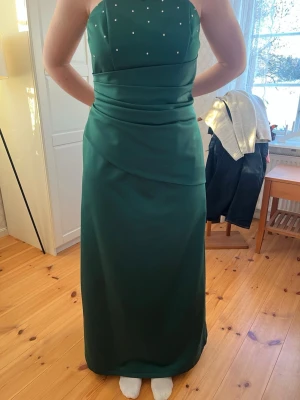 Grön axelbandslös balklänning - Elegant grön axelbandslös balklänning i lång modell med raka linjer och vackra draperingar i midjan. Överdelen är dekorerad med små glittriga pärlor för extra lyxig känsla. Till klänningen hör en matchande sjal och liten väska. Perfekt för bal eller festliga tillfällen. Skickar gärna mått om det skulle viljas, kan även erbjuda provning, bor i Växjö💕 st är 40/42 men hon som bär den på bilden har M i tröja så skulle säga M 💕