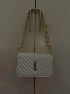 Vit quiltad axelväska från YSL - Säljer en snygg vit axelväska från YSL med guldfärgad kedja och klassisk YSL-logga framtill. Väskan är quiltad i ett elegant mönster och har en stilren, rektangulär form. Perfekt för dig som vill ha en lyxig accessoar med exklusiv känsla.