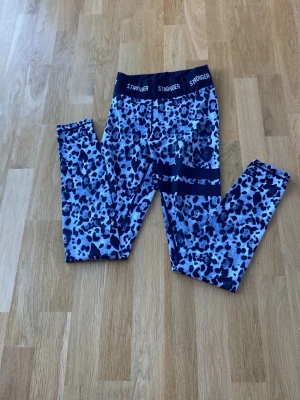 Blå leopardmönstrade leggings Stronger - Säljer ett par snygga blå leggings från Stronger med leopardmönster och bred resår i midjan där det står 'STRONGER'. Perfekta för träning eller chill, stretchigt material som sitter skönt och följer kroppen.