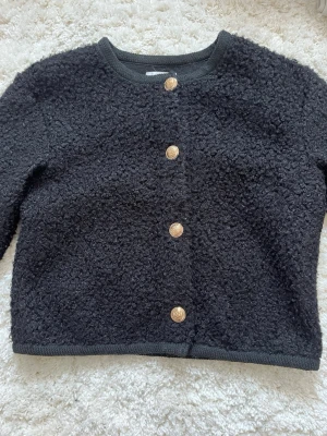 En super mysig svart jacka/kofta - Kort, svart jacka/kofta i boucléliknande material med dekorativa guldknappar framtill. Modellen har rund halsringning, långa ärmar och en något boxig passform som ger ett elegant och trendigt uttryck. Perfekt att bära både till vardags och vid mer uppklädda tillfällen. En mångsidig favorit som passar lika bra till jeans som över klänning eller kjol.❣️❣️