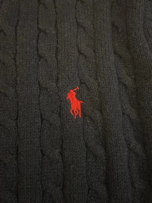 Mörkblå stickad tröja Polo Ralph Lauren - Kabelstickad mörkblå tröja från Polo Ralph Lauren med klassisk röd broderad logga på bröstet. Tröjan har rund halsringning och långa ärmar, perfekt för kyligare dagar. Materialet är mjukt och skönt, och den ribbade kanten ger en snygg passform.