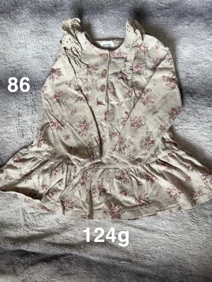 Blommig beige klänning med spets - Söt beige klänning från Newbie med rosa blommönster, spetsdetaljer vid axlarna och knappar framtill. Klänningen har lång ärm och volang nedtill, perfekt för vår och sommar. 🌸Fint använt skick. Storlek 86 