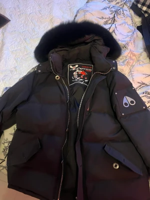 Svart Moose Knuckles pufferjacka - Säljer en svart pufferjacka från Moose Knuckles med stor fluffig huva och pälsdetalj. Jackan har dragkedja, knappar och två stora fickor framtill. Insidan har ett broderat hockeymärke och logga på ärmen. Perfekt för kalla dagar och riktigt snygg streetstil.