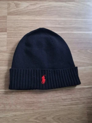 Svart mössa från Polo Ralph Lauren - Klassisk svart mössa från Polo Ralph Lauren med röd broderad logga framtill. Mössan är ribbstickad nedtill och tillverkad i mjuk merinoull som håller dig varm under kalla dagar. Enkel och stilren design som passar till det mesta. Har använt typ 5 gånger, hör av dig om du har frågor.