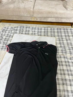 Svart pikétröja med röda detaljer - Säljer en svart pikétröja från B&S med klassisk krage och knappar framtill. Tröjan har röda och vita detaljer på krage och ärmslut som ger en sportig vibe. Perfekt till jeans eller shorts för en clean look.