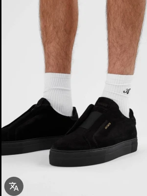 Svarta sneakers från Axel Arigato - Säljer ett par svarta sneakers från Axel Arigato i mocka med minimalistisk design och guldfärgad logga på sidan. Skorna har platt sula och elastisk detalj över vristen för enkel påtagning. Perfekta för dig som gillar stilrena och cleana skor.