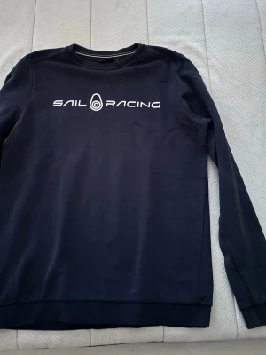 Mörkblå Sail Racing sweatshirt - Snygg mörkblå sweatshirt från Sail Racing med vit logga tryckt på bröstet och en mindre logga i nacken. Klassisk rund halsringning och långa ärmar. Perfekt för dig som gillar stilren design och vill ha något bekvämt till vardags. Knappt andvänd