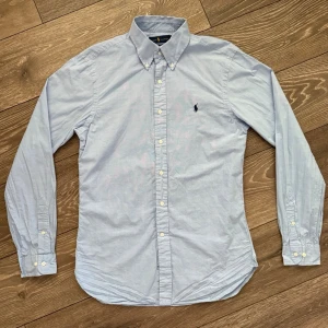 Ralph Lauren skjorta - Snygg rutig ljusblå skjorta från Ralph Lauren i slim fit-modell. Skjortan har klassisk krage med knappar, lång ärm och broderad logga på bröstet. Materialet är mjuk bomull med subtilt rutmönster. Perfekt för dig som gillar stilrena och tidlösa plagg.