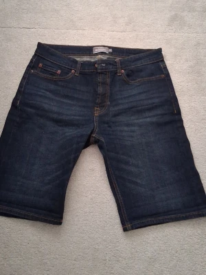 Mörkblå jeansshorts från Dressmann - Säljer ett par mörkblå jeansshorts från Dressmann i klassisk femficksmodell. Shortsen har kontrastsömmar i orange och normal passform. Perfekta för dig som gillar enkel och stilren denim-look.