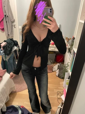 Svart croppad topp med knyt - Säljer en svart croppad topp med lång ärm och djup v-ringning. Toppen har en snygg knytning framtill som ger en feminin touch. Perfekt till jeans eller kjol för en trendig look.