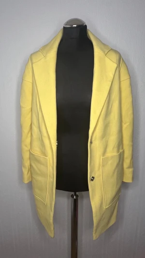 Gul kappa, storlek 36 - Snygg ljusgul kappa med stora fickor och klassisk krage. Rak passform och enkelknäppning framtill. Perfekt för dig som vill sticka ut med färg i vår eller höst. Materialet känns mjukt och har en ullig struktur.