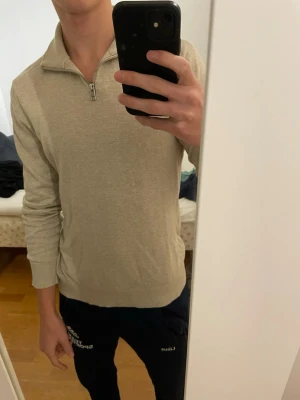 Beige half zip-tröja Jack & Jones - Säljer en stilren beige långärmad tröja från Jack & Jones med half zip-dragkedja i halsen. Tröjan har ribbade muddar och är i mjuk bomullsmix, perfekt för lager-på-lager under hösten eller våren.