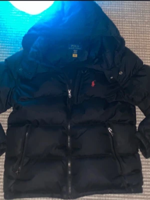 Svart pufferjacka från Polo Ralph Lauren - Svart pufferjacka från Polo Ralph Lauren med huva och två stora fickor med dragkedja. Jackan har ett rött Polo-logotryck på bröstet och quiltad design. Perfekt för kalla dagar och har en riktigt snygg Grisch - stil 