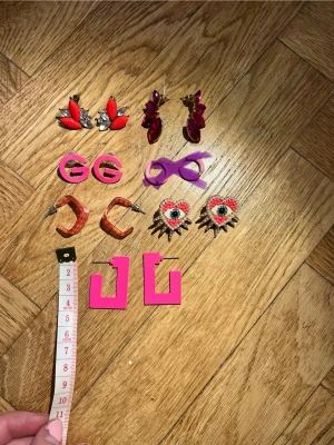 Färgglatt örhängespaket med 6 par - Säljer ett paket med 6 par örhängen i olika former och färger. Inkluderar röda och rosa stenar, rosa GG-loggor, ljusrosa hoops, orange transparenta hoops, hjärtformade örhängen med pärlor och ögonmotiv samt stora neonrosa fyrkantiga örhängen. Perfekt för dig som gillar att sticka ut!