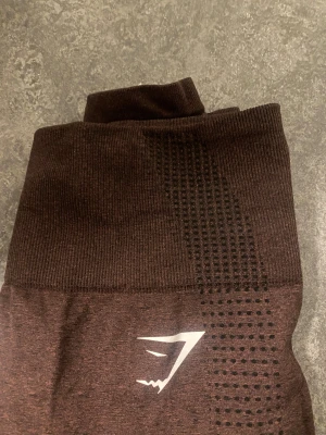 Bruna seamless leggings från Gymshark - Mörkbruna seamless leggings från Gymshark med hög midja och ribbade detaljer längs benen. Snyggt mönstrade meshpaneler på sidorna och logga på låret. Stretchigt material som sitter tight och följer kroppen perfekt.