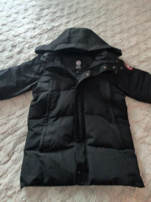 Svart Canada Goose jacka  - Säljer en svart dunjacka från Canada Goose med huva och klassisk patch på ärmen. Jackan har dragkedja och knappar framtill, två stora fickor och quiltad design. Perfekt för kalla dagar och riktigt snygg till vinterlooken. 1 knapp som saknas och 1 som är lite paj och en av dragkedjorna går att öppna halvvägs bara 