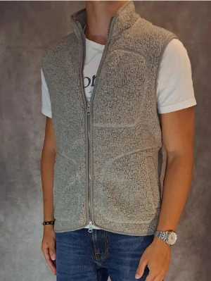 Grå teddyväst från J.Lindeberg - Säljer en grå teddyväst från J.Lindeberg med hel dragkedja framtill och två stora fickor. Västen är gjord i mjukt fleece-material och har en hög krage. Perfekt att slänga över en hoodie eller t-shirt för extra värme.