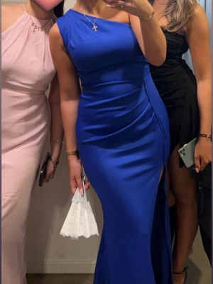 Blå långklänning med slits - Säljer en elegant blå långklänning med oneshoulder och hög slits på sidan. Klänningen har en figurnära passform och är tillverkad i ett slätt, glansigt material som ger en lyxig känsla. Perfekt för dig som vill sticka ut med en stilren och modern look. Vet inte storlek men antingen S eller M