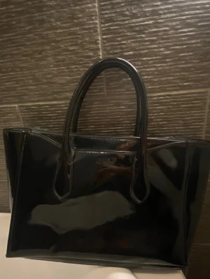 Svart handväska från POLO Ralph Lauren  - Säljer en stor och glansig svart handväska från POLO Ralph Lauren i lackat skinn. Väskan har två rejäla handtag och en avtagbar axelrem. Väskan är aldrig använd så den är precis som ny. Går ej att hitta i butik 
