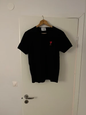 Svart t-shirt från Ami Paris - Svart t-shirt från Ami Paris med klassisk passform och korta ärmar. På bröstet finns ett broderat rött hjärta med bokstaven A. Tillverkad i mjuk bomull och perfekt för en clean och stilren look.
