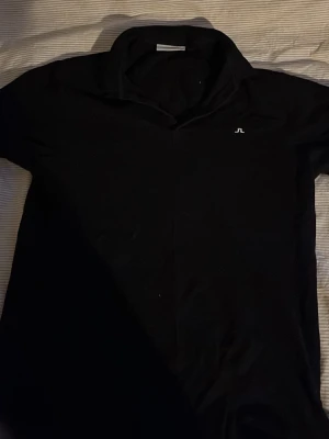 Svart piké från J.Lindeberg - Klassisk svart pikétröja från J.Lindeberg med liten vit logga på bröstet. Skön passform och mjukt material, perfekt till jeans eller shorts. Snygg krage och korta ärmar ger en clean look. Skicka bud hur mycket ni är villiga att köpa tröjan för