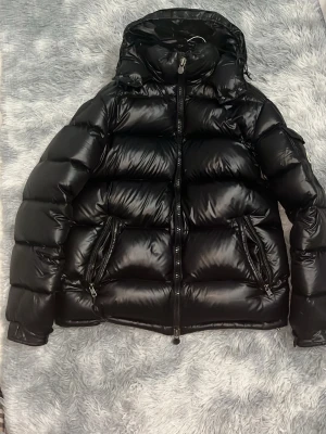 Svart pufferjacka från Moncler - Säljer en svart pufferjacka från Moncler med glansig finish och huva. Jackan har två stora fickor med dragkedja och klassisk Moncler-logga på ärmen. Fodrad med dun och fjäder för extra värme, perfekt för kalla dagar. Stängs med dragkedja framtill.